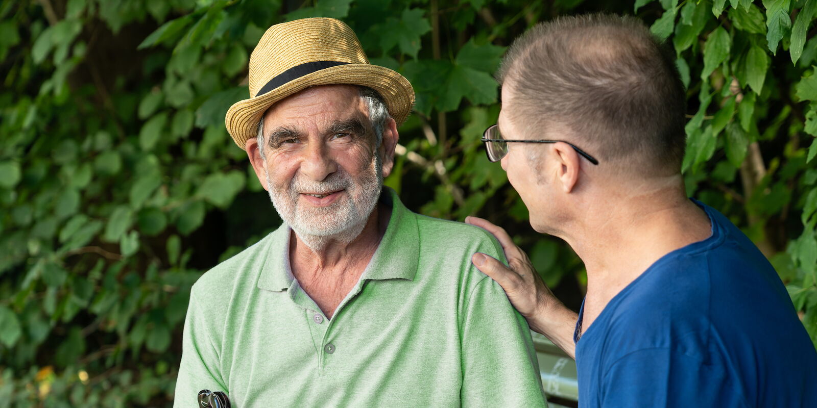 Ein älterer Mann sitzt auf einer Bank; neben ihm sitzt eine Betreuungsperson.