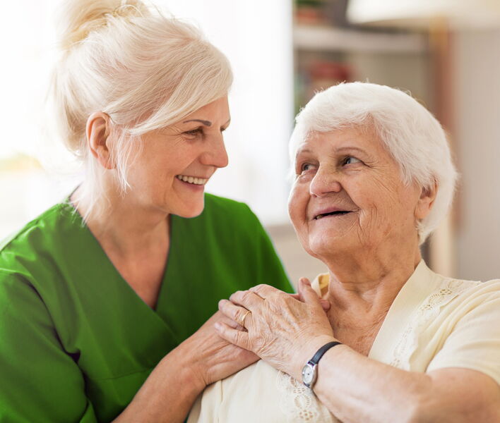 Eine Betreuungsperson sitzt neben einer älteren Frau, hält ihre Hand und lächelt sie herzlich an.