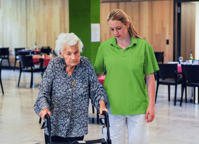 Eine ältere Frau geht mit einem Rollator durch einen Raum, begleitet von einer Betreuungsperson an ihrer Seite.