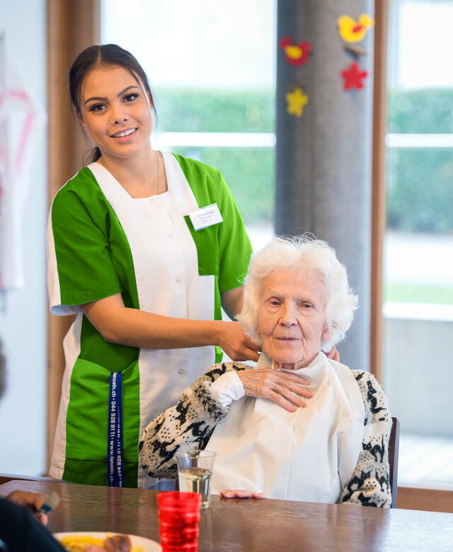 Eine Pflegefachperson steht hinter einer älteren Frau am Tisch während der Essenszeit.
