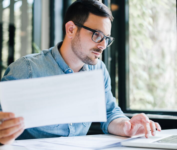 Eine Person sitzt am Tisch, arbeitet am Laptop und hält dabei ein Dokument in der Hand.